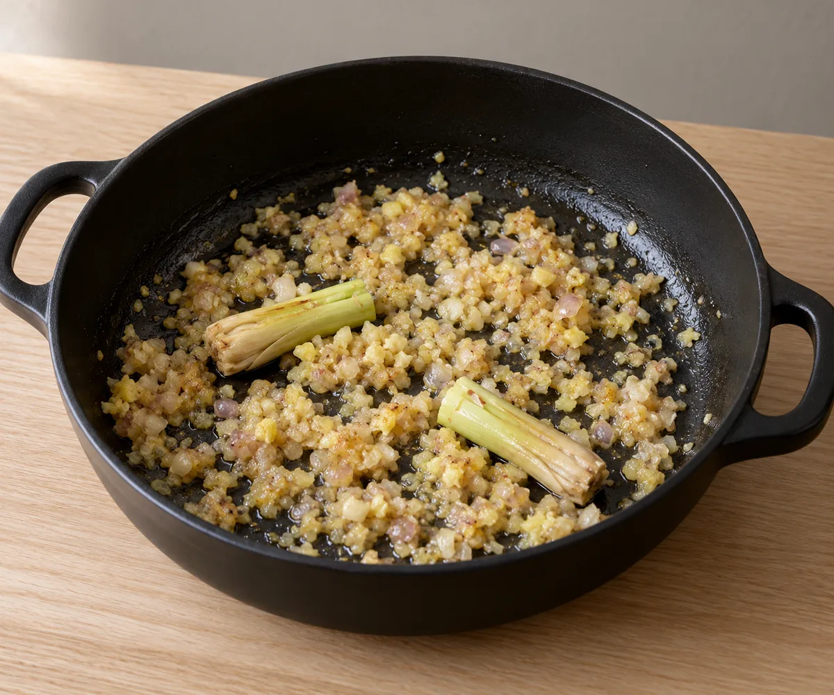 Finely chopped aromatics softened to pale gold with two short lemongrass pieces in a black casserole.